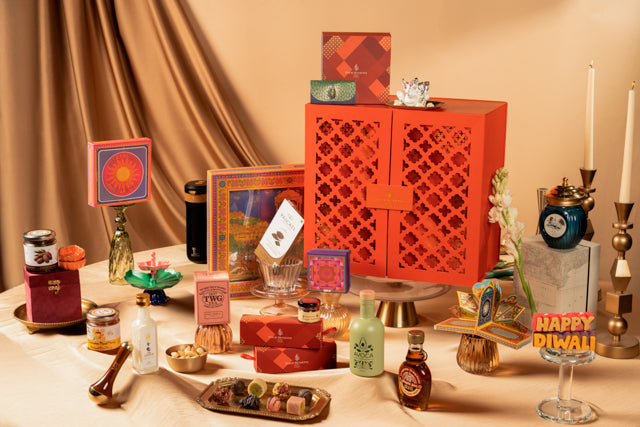 A styled Diwali gift table display featuring a prominent red-orange lattice-patterned cabinet, surrounded by assorted decorative items including framed art, candles, a blue teapot, small bottles, and packaged treats on a draped beige fabric background.