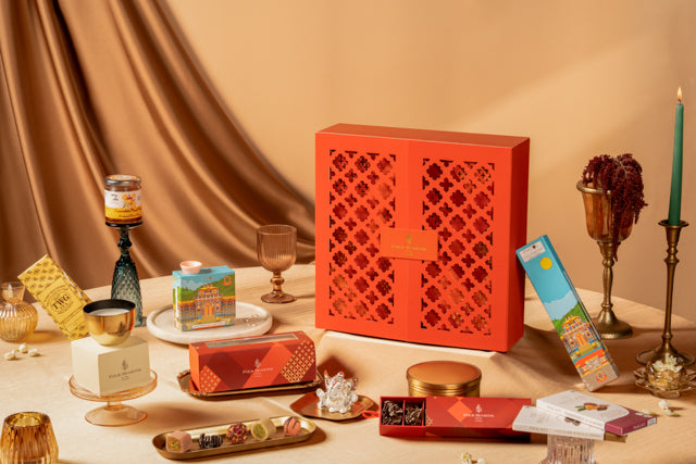 A luxury Diwali gift set featuring a prominent red-orange double-door box with intricate laser-cut geometric patterns, displayed on a table surrounded by gourmet chocolates, tea, and decorative items against a draped beige background.