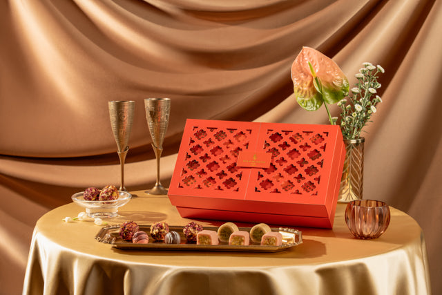 A red rectangular Diwali gift box with a lattice cutout lid sits on a draped champagne tablecloth, surrounded by chocolate pieces, two champagne flutes, a small bowl, and decorative flowers against a matching satin backdrop.
