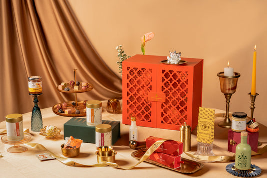 A styled product display featuring a prominent orange-red lattice-patterned decorative box, surrounded by candles, jars, and gift boxes on a draped beige surface with a warm brown curtain background.