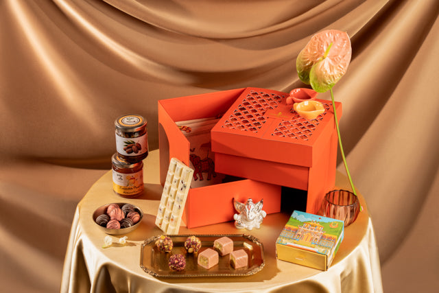 An orange gift box set displayed on a draped champagne tablecloth with assorted chocolates, jars, and a small decorative box. The scene is styled with warm lighting and a satin fabric backdrop.