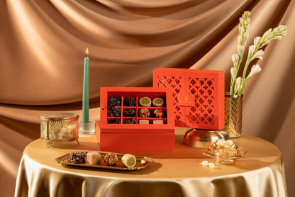 A luxury red Diwali gift box set of assorted chocolates and sweets, styled on a draped champagne tablecloth with a teal candle, gold dishes, and white flowers against a satin gold backdrop.