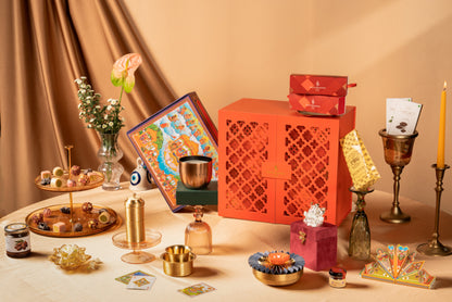 A styled tabletop arrangement of decorative objects in warm neutral tones, including a bright orange carved storage box, a colorful illustrated box, brass candlesticks, a yellow taper candle, small gift boxes, glass bottles, and floral accents against a draped beige backdrop.
